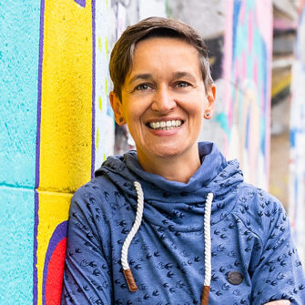 Barbara Andrä lehnt im freien an an der bunten Wand und lächelt in die Kamera.